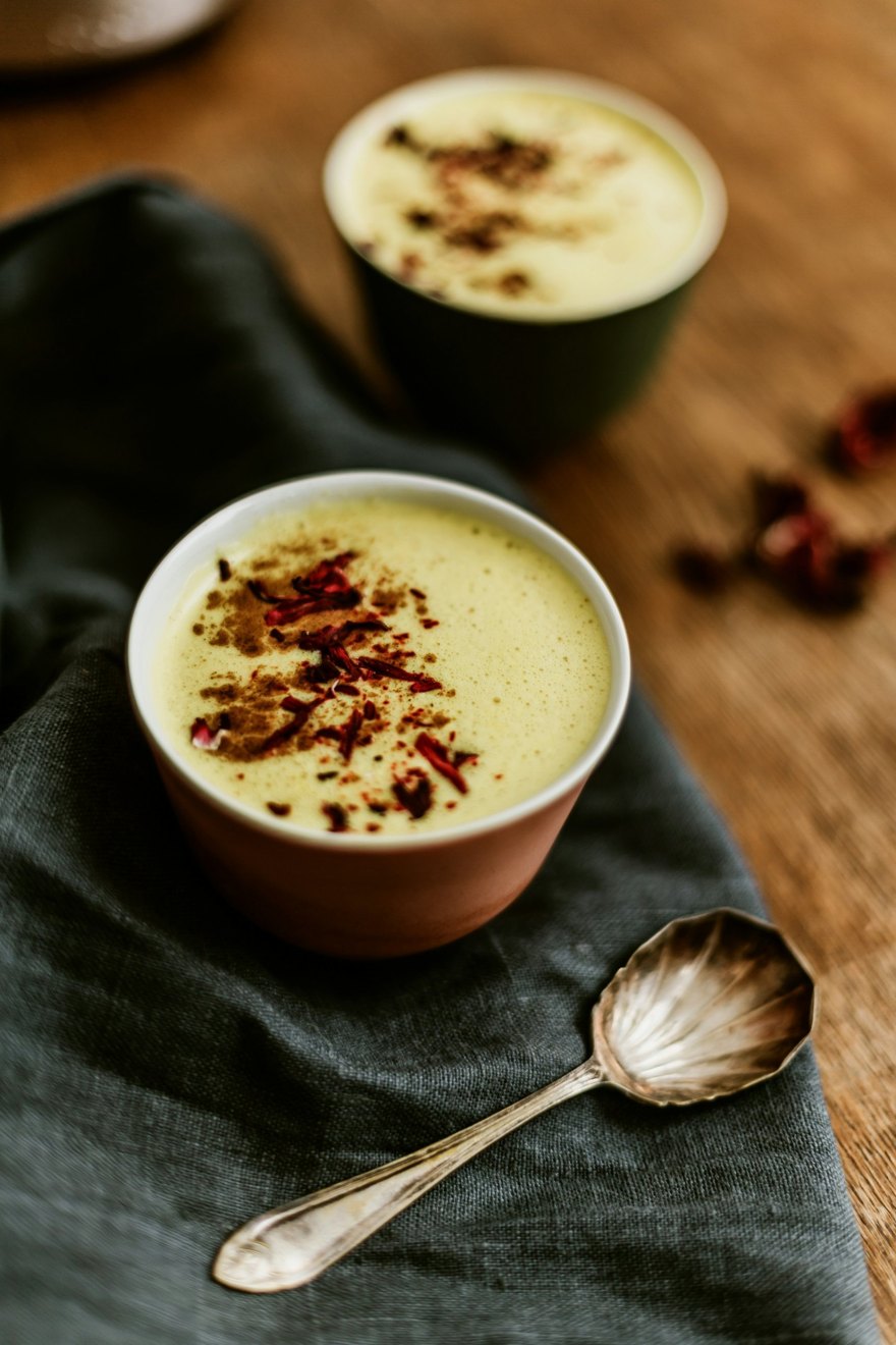 A cup of frothy golden milk dusted with cinnamon and dried flower petals, with a vintage spoon on a linen napkin